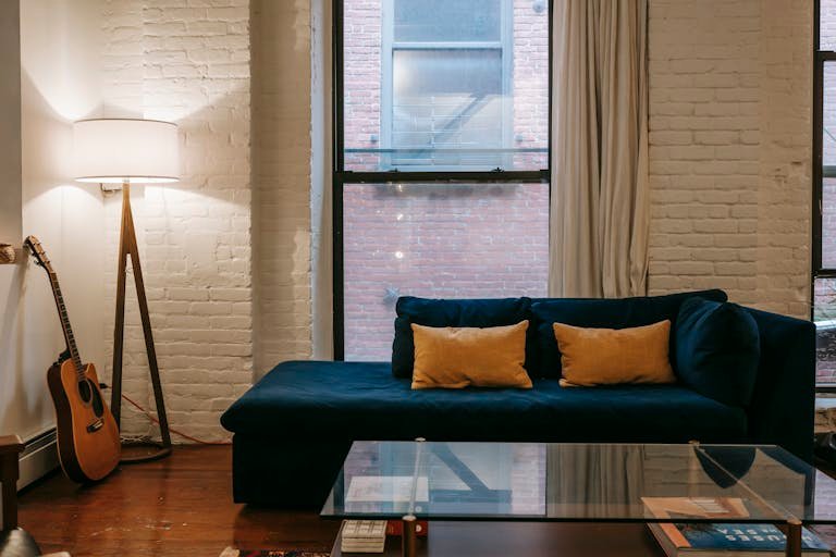 Comfortable blue sofa with yellow cushions placed near white brick wall and guitar with lamp in cozy living room with glass table